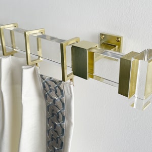 Peut inclure: Tringle à rideaux dorée avec des accents en acrylique transparent et des embouts carrés. La tringle est fixée à un mur blanc et des rideaux blancs y sont suspendus.