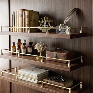May include: Three dark wood wall shelves with brass railings, each holding various items including vintage books, a brass sextant, a glass inkwell with a quill, amber apothecary bottles, a white coral sculpture, a leather box, and metal keys.