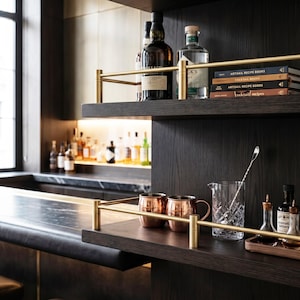 Könnte beinhalten: Dunkle Holzregale mit Messinggeländern, die Glasflaschen, Cocktailbücher, Kupferbecher, ein Kristall-Rührglas mit langem Rührlöffel sowie kleine Glasflaschen mit Bitter auf einer Bar-Thekenoberfläche präsentieren.