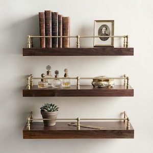 May include: Three horizontal dark wood wall shelves with brass railings. The top shelf holds five vintage books and a framed portrait. The middle shelf features three glass perfume bottles and a decorative box. The bottom shelf holds a succulent.