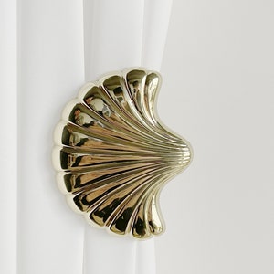May include: Gold-toned seashell curtain tieback. The tieback is shaped like a seashell with a smooth, shiny finish.