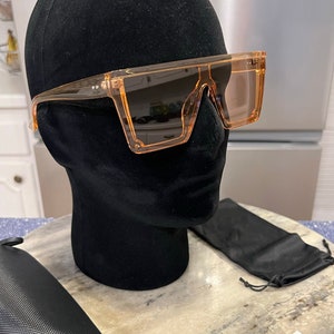 May include: Orange tinted square sunglasses with a black case and a black drawstring pouch. The sunglasses are on a black mannequin head. The text "PERFECT PACKING" is on a white label.