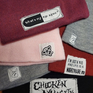 May include: A collection of six knit beanies in various colors including maroon, light pink, grey, red, and black. Each beanie features a rectangular or square fabric patch with graphic designs or text, including phrases like What's my age again? and I'm just a kid and life is a nightmare, plus hand gesture and flower icons.