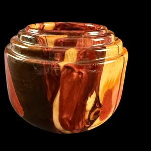 May include: A handcrafted wooden bowl with a unique design. The bowl features a layered, tiered rim and a rounded body. The wood grain showcases a blend of dark brown, red, and light tan hues, creating a striking visual contrast. The bowl is set against a black background.
