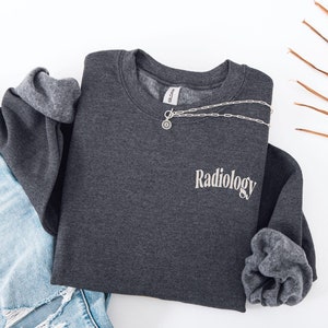 May include: Dark heather grey sweatshirt with the word "Radiology" embroidered on the front in a white script font.
