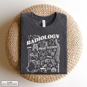 以下が含まれることがあります： ダークヘザーグレーのTシャツに、放射線機器と「RADIOLOGY」というテキストの白いグラフィックデザインが施されています。グラフィックには、骨格、X線、フィルムカセット、放射線記号、その他の医療機器が含まれています。