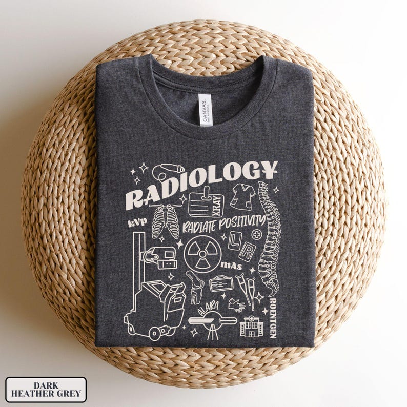 以下が含まれることがあります： ダークヘザーグレーのTシャツに、放射線機器と「RADIOLOGY」というテキストの白いグラフィックデザインが施されています。グラフィックには、骨格、X線、フィルムカセット、放射線記号、その他の医療機器が含まれています。