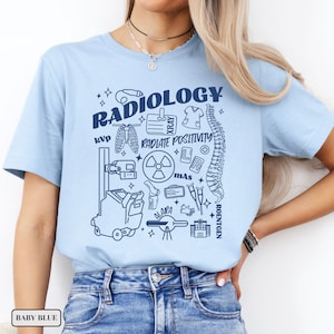 以下が含まれることがあります： 放射線学の様々なシンボルと「RADIOLOGY RADIATE POSITIVITY」というテキストが描かれたライトブルーのTシャツです。