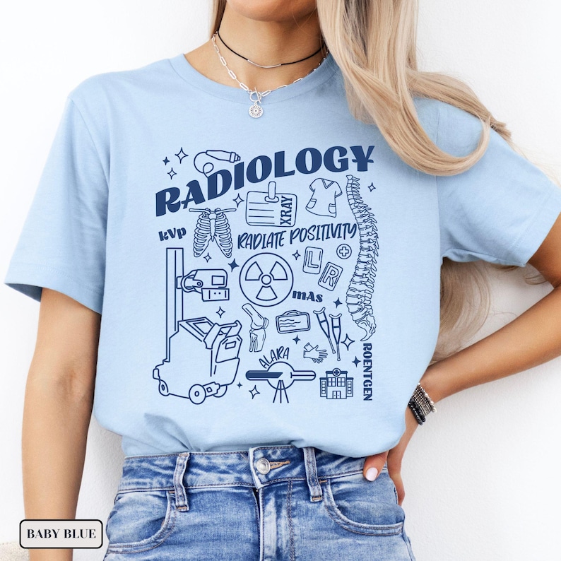 以下が含まれることがあります： 放射線学の様々なシンボルと「RADIOLOGY RADIATE POSITIVITY」というテキストが描かれたライトブルーのTシャツです。