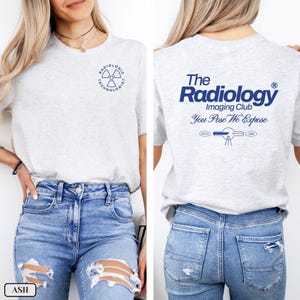 May include: A heather grey t-shirt with a blue graphic of a radiation symbol and the text "Radiologic Technologist". The back of the shirt has a blue graphic with the text "The Radiology Imaging Club You Pose We Expose" and the text "ESTO 1970 3866".