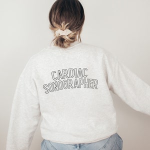 May include: A light gray sweatshirt with the words "CARDIAC SONOGRAPHER" printed in black, outlined letters. The sweatshirt is being worn by a person with their back to the camera. The person has brown hair pulled back in a bun with a beige scrunchie.