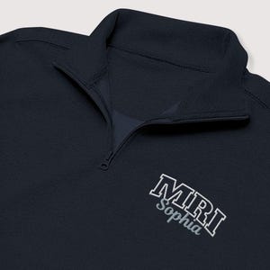 Peut inclure: Polo à quart de fermeture éclair bleu marine avec le texte brodé "MRI Sophia" en blanc et gris. Le pull-over a un col montant et une fermeture éclair qui descend sur la poitrine. Le tissu a un aspect texturé.