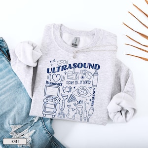Peut inclure: Un sweat-shirt gris clair avec le mot "ULTRASOUND" en bleu marine, accompagné d'illustrations médicales. Le motif comprend les mots "HARMONICS", "SCAN TIL IT HERTZ" et "ALARA". Le sweat-shirt est présenté avec un jean bleu.