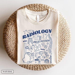 以下が含まれることがあります： ベージュのTシャツに、青いグラフィックデザインが施されています。デザインには「Radiology」の文字と、レントゲン写真、骨格、聴診器など、さまざまな医療イラストが描かれています。「Radiate Positivity」という文字もデザインに含まれています。