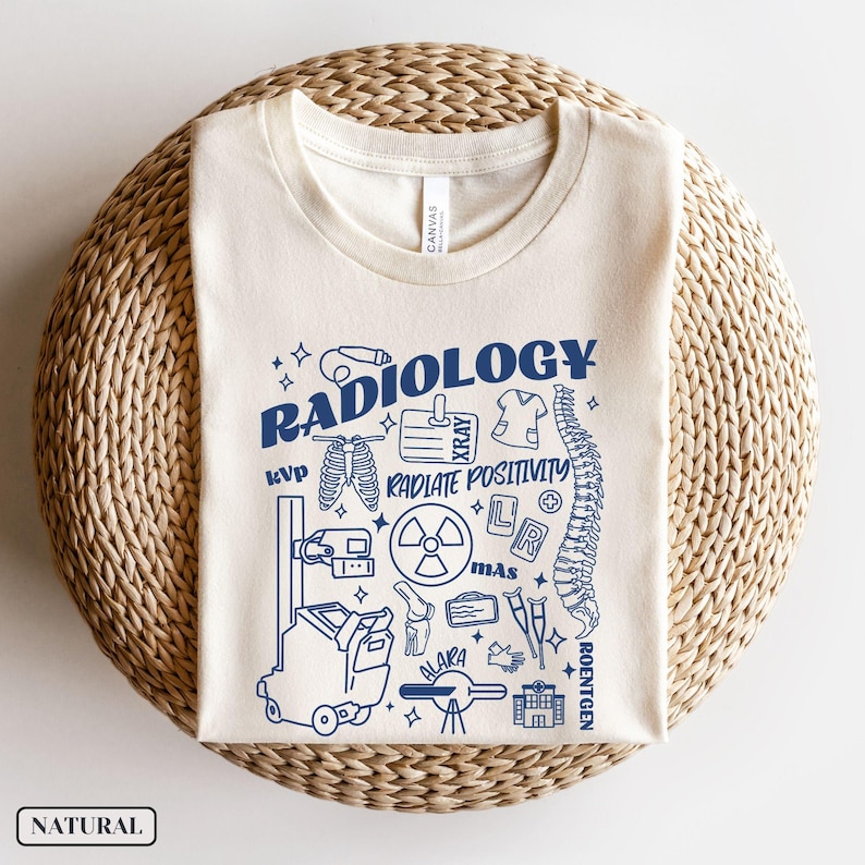 以下が含まれることがあります： ベージュのTシャツに、青いグラフィックデザインが施されています。デザインには「Radiology」の文字と、レントゲン写真、骨格、聴診器など、さまざまな医療イラストが描かれています。「Radiate Positivity」という文字もデザインに含まれています。