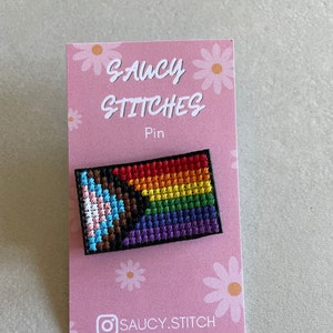 May include: A rainbow pride flag with black and brown stripes is stitched on a pin with a black border. The pin is on a pink card with white daisies and the text "SAUCY STITCHES Pin" and "@SAUCY.STITCH".