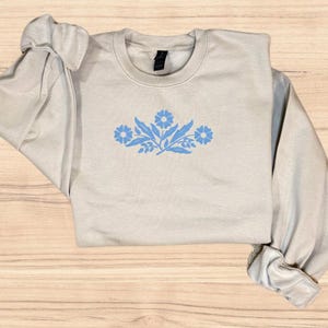 May include: A light beige sweatshirt with a blue floral design on the front. The design features stylised flowers and leaves.
