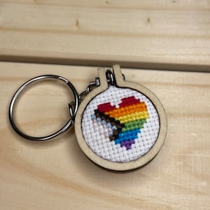 May include: A rainbow heart-shaped design is embroidered on a small white canvas, framed in a wooden circle. The canvas is attached to a silver key ring.