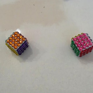 May include: Two cube-shaped dice covered in small, multi-colored rhinestones. The left die features orange, purple, and yellow stones, while the right die displays pink, green, and red stones. Each face of the dice is fully encrusted with gems.
