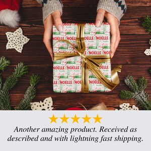 Puede incluir: Una caja de regalo envuelta con una cinta dorada, sostenida por manos. El papel de regalo es blanco con texto rojo y verde que dice "NOELLE". La caja est&aacute; rodeada de decoraciones navide&ntilde;as sobre una superficie de madera.