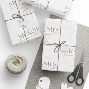May include: White wrapping paper with a marble pattern, printed with the text Mr & Mrs, James and Amanda Wilson, and the date June 10, 2025. The paper features line art of two rings and is tied with brown twine and white star or heart tags.