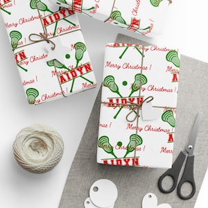 May include: White wrapping paper with a repeating pattern of green lacrosse sticks and the text "Merry Christmas!" in red. The wrapped gifts are tied with twine and have small white tags. A ball of twine and scissors are also visible.