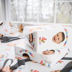 May include: White wrapping paper featuring a repeating pattern of a child's face, the text "HAPPY BIRTHDAY", a red number "4", and a small green dinosaur graphic. The paper is used to wrap two rectangular boxes, with a black ribbon and scissors nearby.