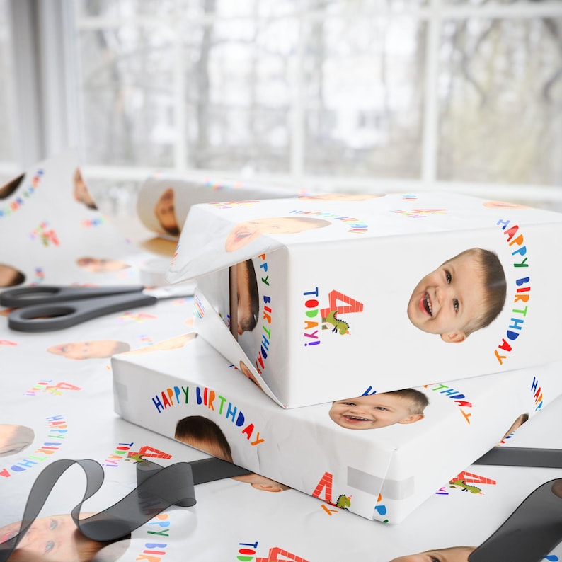 May include: White wrapping paper featuring a repeating pattern of a child's face, the text "HAPPY BIRTHDAY", a red number "4", and a small green dinosaur graphic. The paper is used to wrap two rectangular boxes, with a black ribbon and scissors nearby.
