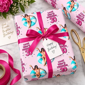 May include: A rectangular gift box wrapped in light pink paper featuring a repeating pattern of a person in a bikini on a blue and white pool float with the text Look Who's Turning 30. The box is tied with a dark pink ribbon and a gift tag.