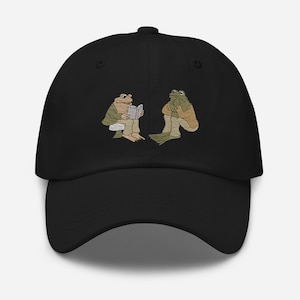 May include: Black baseball cap with an embroidered design of two frogs sitting and reading a book.