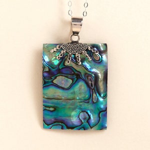 Puede incluir: Un collar con colgante rectangular con un diseño de concha de abulón brillante en tonos verdes, azules y morados. El colgante está sujeto a una cadena de plata. La parte superior del colgante tiene un elemento decorativo plateado.