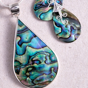 May include: Abalone shell jewelry set featuring a pendant and matching earrings. The teardrop-shaped pieces display iridescent blues, greens, and purples, set in silver. The earrings have decorative silver swirls.