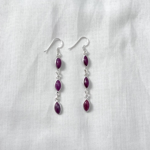 May include: A pair of drop earrings featuring three oval, deep red gemstones set in silver-coloured metal. The earrings hang from a simple hook design. The background is a white, textured fabric.