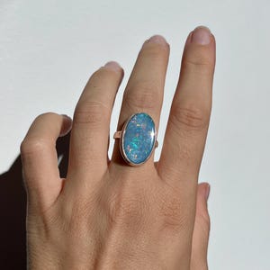 May include: An oval opal ring with a silver band. The opal displays blue and iridescent colours, with flashes of green and purple. The ring is set on a hand against a white background. A piece of jewellery.