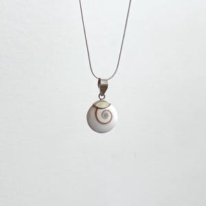 Puede incluir: Un collar plateado con un colgante blanco con un diseño de espiral marrón. El colgante redondo cuelga de una delicada cadena plateada. El fondo es blanco liso.