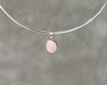 May include: A silver-toned choker necklace with a small, oval, light pink pendant. The pendant is a smooth, polished stone. The necklace is displayed against a neutral gray background.