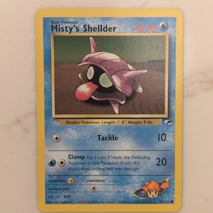 May include: A Misty's Shellder Pokemon card from the 1999-2000 Wizards set. The card is yellow with a blue border and features an illustration of a purple and pink Shellder Pokemon. The card has a blue energy symbol in the top right corner and a white star in the bottom left corner. The card's name is "Misty's Shellder" and its HP is 40. The card has two attacks: "Tackle" for 10 damage and "Clamp" for 20 damage. The card's weakness is electric and its retreat cost is one energy card. The card is number 89/132.