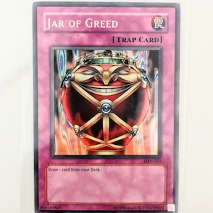 Può includere: Una carta da gioco "Jar of Greed" rossa e dorata del gioco di carte collezionabili Yu-Gi-Oh! La carta presenta un'illustrazione di un barattolo rosso sorridente con un coperchio dorato e una cornice dorata. Il testo della carta recita "Pesca 1 carta dal tuo Deck."