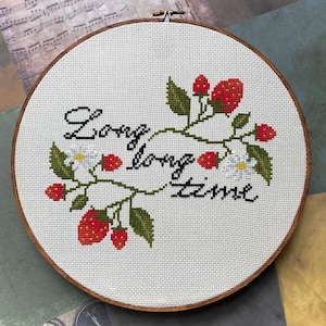 May include: A white embroidery hoop with a cross-stitch design of a green vine with red strawberries and white flowers. The text "Long long time" is stitched in black thread.