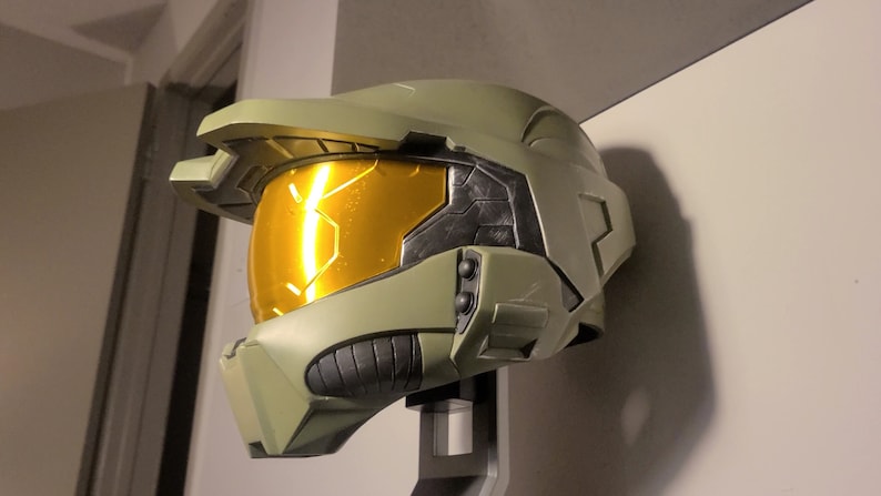 May include: A green and black plastic replica of a Master Chief helmet from the Halo video game series. The helmet has a gold visor and is mounted on a stand.