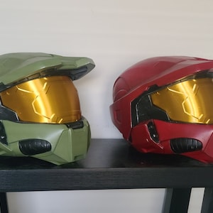 May include: Two plastic helmets, one green and one red, with gold visors. The helmets are sitting on a black shelf.