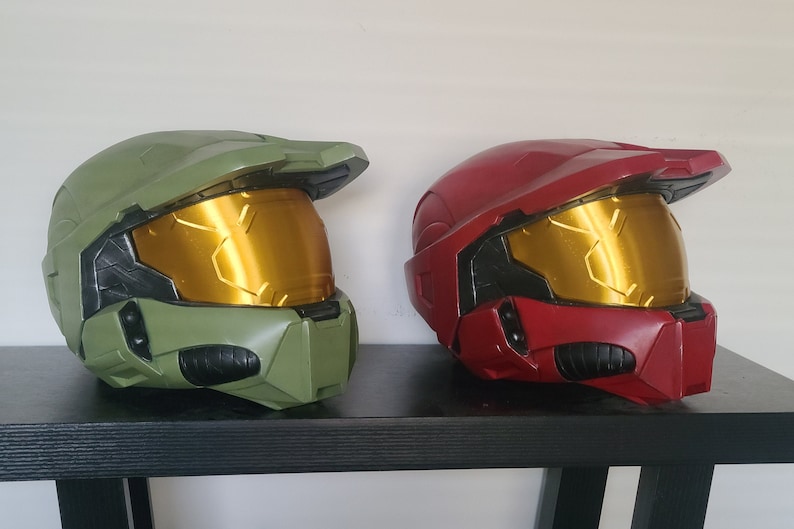 May include: Two plastic helmets, one green and one red, with gold visors. The helmets are sitting on a black shelf.