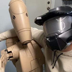 May include: A person wearing a black and gray helmet with a silver visor is standing next to a beige robot with a round head and a long, thin body. The robot has a series of joints and appears to be made of plastic.