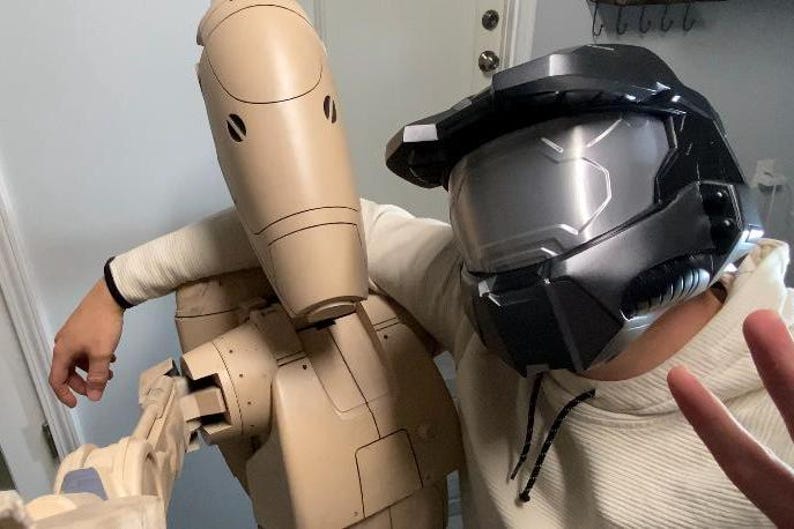 May include: A person wearing a black and gray helmet with a silver visor is standing next to a beige robot with a round head and a long, thin body. The robot has a series of joints and appears to be made of plastic.