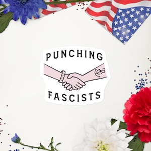Puede incluir: Una pegatina blanca con el texto "PUNCHING FASCISTS" en negro, sobre una ilustración de dos manos estrechándose. Una mano tiene una pulsera, la otra un tatuaje de ancla. La pegatina está rodeada de decoraciones rojas, blancas y azules.