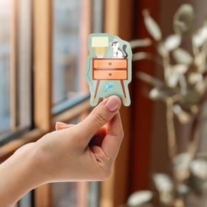 Könnte beinhalten: Ein Aufkleber mit einer Cartoon-Illustration eines Nachttisches mit einer Lampe und einer Katze. Der Nachttisch hat zwei orangefarbene Schubladen und eine hellgrüne Basis. Die Katze ist schwarz und weiß. Der Aufkleber wird von einer Person gehalten.