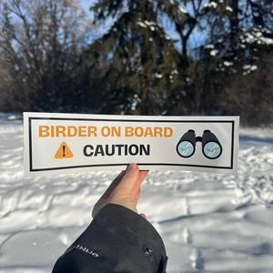 May include: A white sticker with black text and a black and white illustration of binoculars. The sticker reads "Birder on Board" with a yellow exclamation point and the word "CAUTION" below.