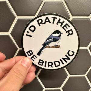 May include: A circular sticker featuring the phrase "I'd Rather Be Birding" around a black-capped chickadee illustration. The bird is depicted in black, white, and grey. The sticker is held against a dark grey hexagonal tile background.