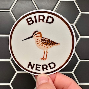 May include: A circular sticker with a brown border, featuring the words "BIRD NERD" in brown. The sticker shows a cartoon bird with brown and tan feathers, a long black beak, and orange legs.
