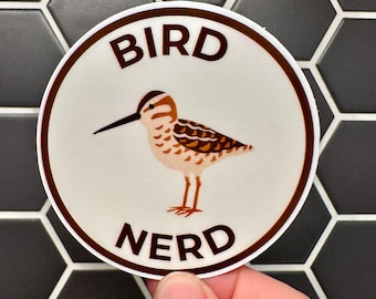 Pode incluir: Um autocolante redondo com uma borda castanha e as palavras "BIRD NERD" em texto castanho. O autocolante apresenta uma ilustração de desenho animado de um pássaro com penas castanhas e bege, um longo bico preto e pernas laranja.